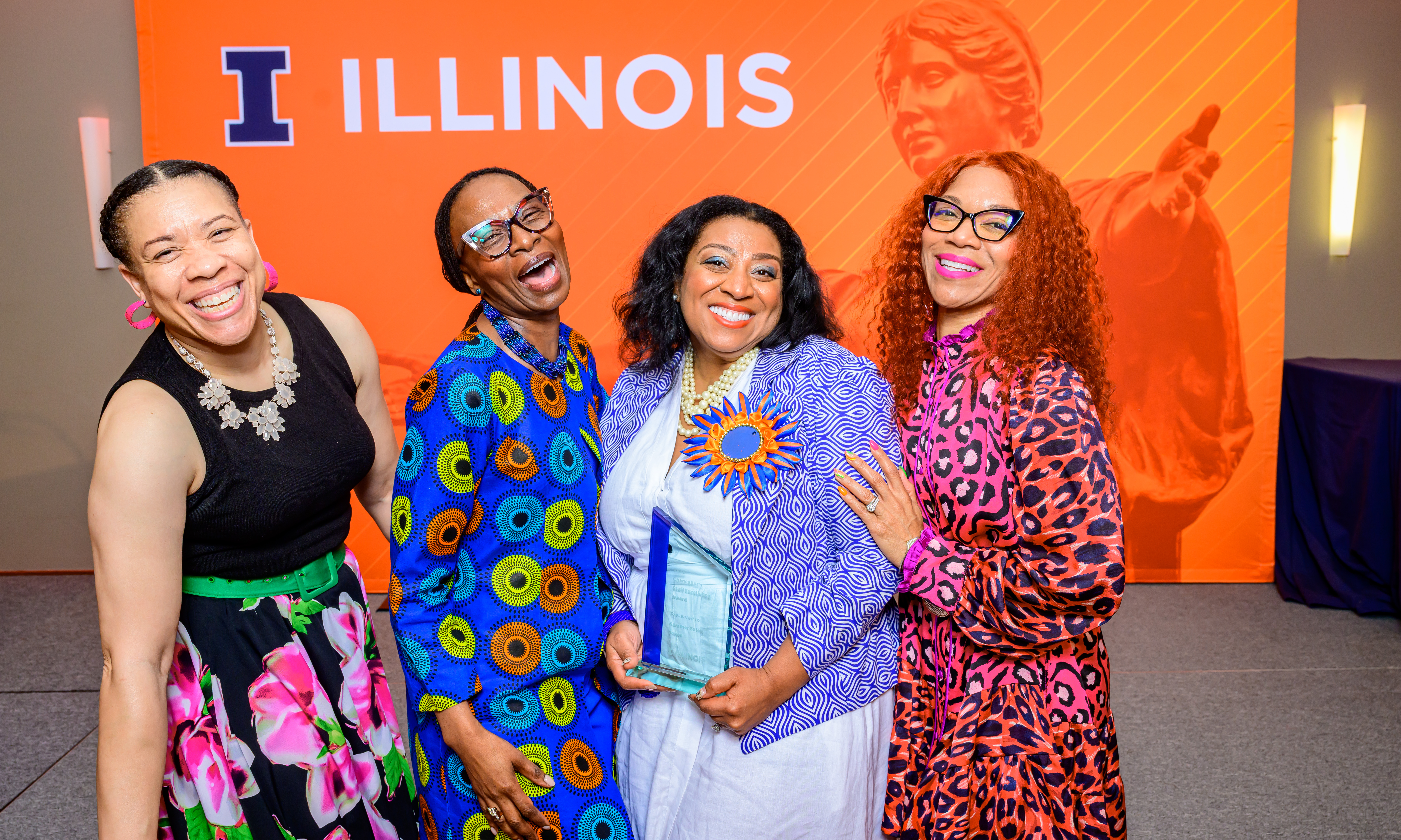 Four people in colorful outfits pose together at Chancellor’s Staff Excellence Award (CSEA) ceremony, with one person holding the CSEA glass award in front of an orange “ILLINOIS” backdrop.